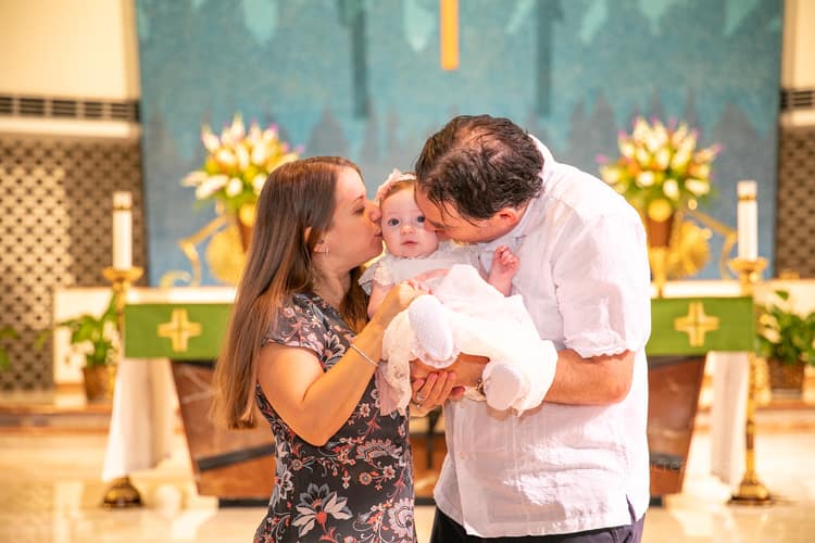 Mother gently holding her baby dressed in white baptism gown inside St. Rose of Lima Church in Miami